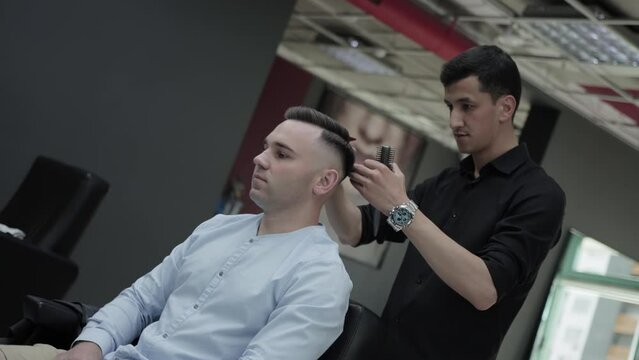 A handsome male hairdresser combs the hair of a young caucasian male client with a comb after a haircut. Professional fashionable haircut in a barbershop. Slow motion