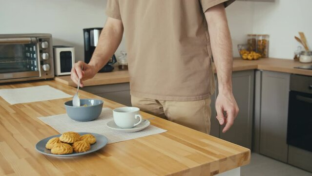 Midsection side stab footage of unrecognizable man serving breakfast coffee and bowl of cereal placing them to light placemat on wooden table near plate of cookies in modern kitchen