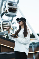 A pretty woman in a black cap eats sushi against the background of the Ferris wheel and the mountains. Sushi concept