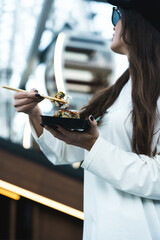 A pretty woman in a black cap eats sushi against the background of the Ferris wheel and the mountains. Sushi concept