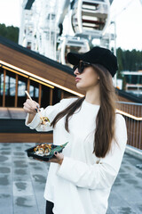 A pretty woman in a black cap eats sushi against the background of the Ferris wheel and the...