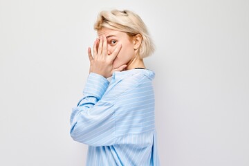 Portrait of young blonde woman covering eyes with hand, peeking through fingers isolated on white...