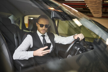 Fototapeta na wymiar handsome charming businessman in black vest with sunglasses looking at phone behind steering wheel