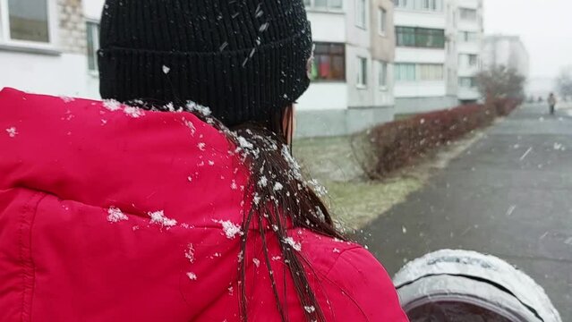 Flakes of wet snow fall on the red hood of a girl who is walking with a stroller in the city. First snow, walking