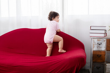 Portrait Multiethnic Hispanic and Asian toddler girl infant climbing red sofa at home looking to window, happy healthy cute baby kid soft skin waiting for mother or playing alone, accident caution