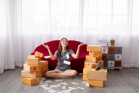 Happy freelance Asian female working selling clothes online sitting with many cardboard boxes at home with full of banknotes falling or flying down to hands, successful small business entrepreneur