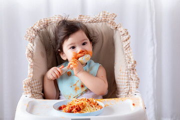 Portrait of happy little toddler infant girl  full of sauce over mouth and face enjoying eating spaghetti, close-up cute baby girl holding spoon try to feed herself in baby seat at home looking camera