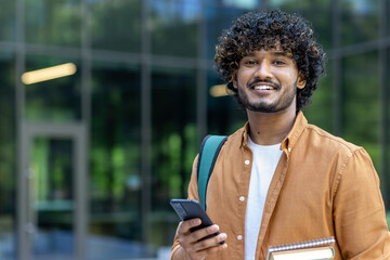Close-up portrait of young smiling Muslim male student holding books and mobile phone and looking...