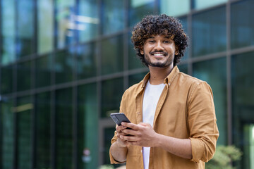 Portrait of a happy and smiling young Muslim man standing on a city street using a mobile phone and...