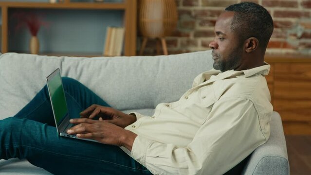 African American man male businessman freelancer lying couch in living room typing on laptop browsing media network make purchase online send email message using computer app relaxing on sofa at home
