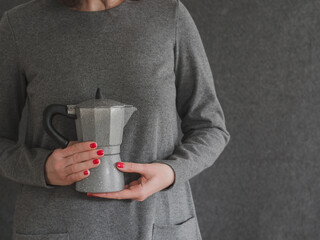 Close-up of a coffee maker held in the hands of a woman dressed in grey against a grey background
