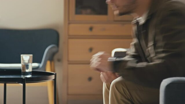 Cropped side stab footage of unrecognizable man reading psychology book sitting on sofa and leaving after checking time on watch in modern living room