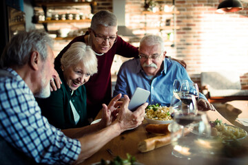 Group of senior friends enjoying a meal together while using smartphone at home