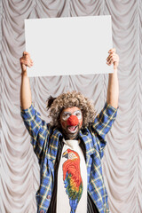 A funny clown shows an empty white sign for the inscription. A clown stands on the stage against the backdrop of the stage scenes