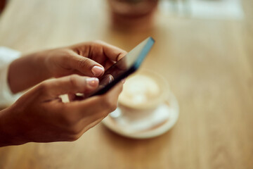 Close-up of a female hands, using a mobile phone, a cup of coffee in the background. - obrazy, fototapety, plakaty