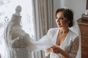 A happy bride is preparing for her luxurious wedding in a hotel room, with a wedding dress on a mannequin nearby. Portrait of a woman with fashionable hair, makeup and a smile in a dressing gown.