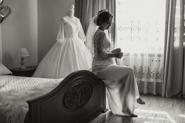 A happy bride is preparing for her luxurious wedding in a hotel room, with a wedding dress on a...
