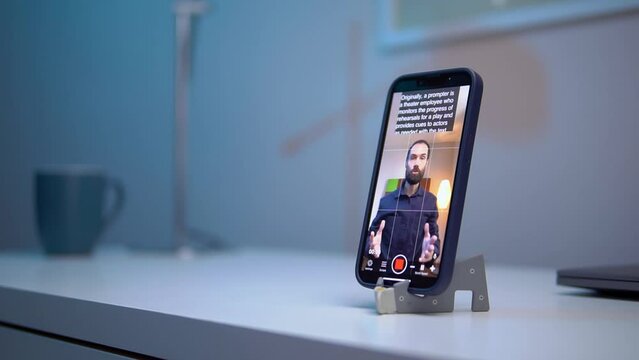 A young man shoots a video on a smartphone while reading text from a teleprompter on a screen