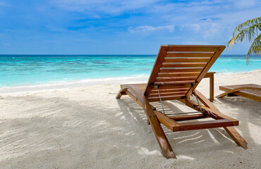 Sun loungers on sandy beach inf front of blue ocean, palm tree leaves framing the scene, Maldives...