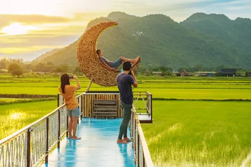 Poster Man using camera to take photo with happiness young couple on crescent moon chair made of rattan in paddy field with beautiful scenic in evening. Decorative wooden moon furniture for viewpoint. © JinnaritT