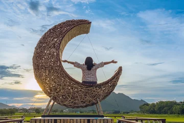 Zelfklevend Fotobehang Woman sit on crescent moon chair made of rattan for relaxation on bridge in paddy field with beautiful scenic in evening. Decorative wooden moon furniture as sitting chair for viewpoint in rice field © JinnaritT