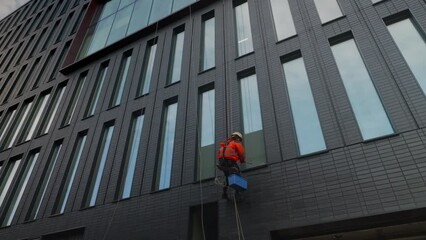 Industrial climber washes windows of a high-rise building, office center complex.A man suspended on ropes with safety gear is doing cleaning service.Occupational hazards, work at height.HQ 4k footage