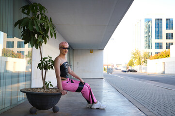 Young gay boy with pink hair and make-up and sunglasses is sitting on a wall resting, in the...