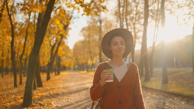 camera treking young stylish woman in autumn city park with yellow foliage against sunset sun with sunflare. attractive girl in hat with paper cup of coffee walks in park along road at warm fall day.