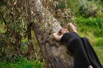 Young woman, blonde and beautiful, with a black dress, with her eyes closed, lying on the trunk of a big tree, in the middle of nature. Concept nature, peace, tranquility, trees.