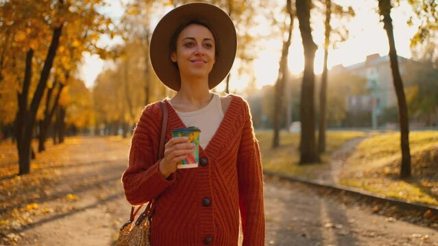 camera treking young stylish woman in autumn city park with yellow foliage against sunset sun with sunflare. attractive girl in hat with paper cup of coffee walks in park along road at warm fall day.