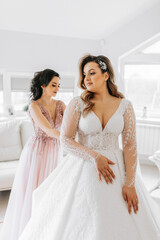 A friend helps the bride fasten her dress. A woman helps her friend fasten the buttons on the back of her wedding dress. The morning of the bride, the creation of a family, an important event.