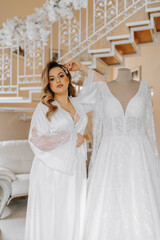 Morning of the bride before the wedding. A beautiful young woman with a veil and a beautiful hairstyle in a white robe near her wedding dress on a mannequin. Professional makeup. Last preparations.