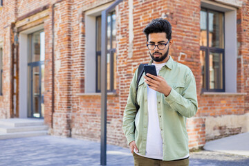 Serious young Indian man standing with backpack on city street, using mobile phone, reading and writing messages, making phone calls.