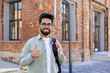 Portrait of a young Indian male student, freelancer standing outside on the street with a backpack,...