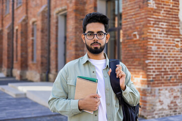 Confident young male Indian student standing outside a university campus with a backpack, holding a...