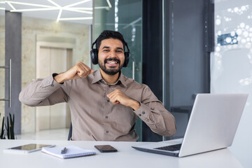 Portrait of a happy young Indian man sitting at a desk in the office, working on a laptop, looking...
