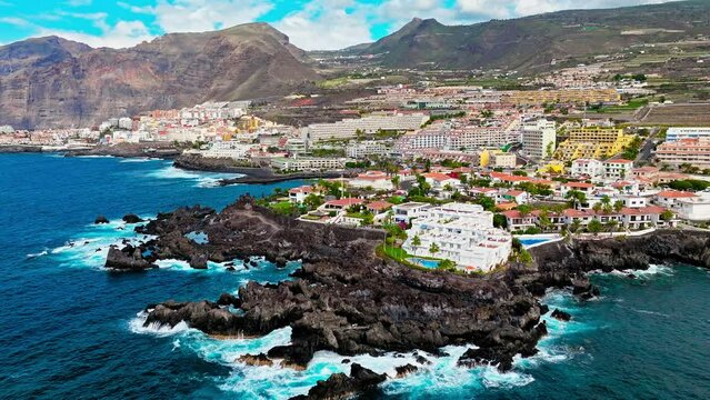 Aerial view of a luxury resort and colourful hotels with swimming pools in Tenerife. Whitewashed houses on a rugged hillside that tumbles down to a tiny black sandy beach in Puerto de Santiago.
