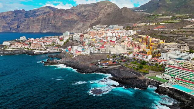 Aerial view of a luxury resort and colourful hotels with swimming pools in Tenerife. Whitewashed houses on a rugged hillside that tumbles down to a tiny black sandy beach in Puerto de Santiago.
