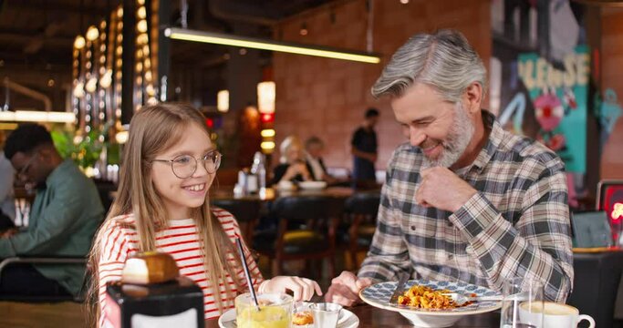 Caring handsome father telling jokes to his little daughter while sitting at table with delicious food and drink. Happy Caucasian family having fun while spending time in cafe or pizzeria.