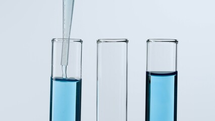 Three glass test tubes on a white background. Two test tubes are filled with blue liquid, a blue substance is dripping into one of them from a pipette. Concept of medicine, biochemical research. Close