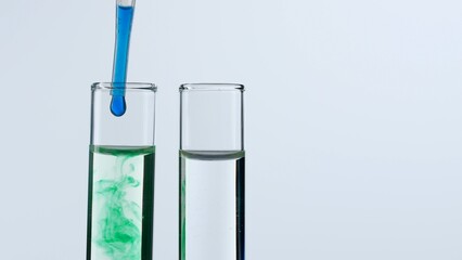 Two glass test tubes on a white background are filled with a transparent liquid. A blue substance drops into one of the test tubes from a pipette and dissolves in green color. Close up