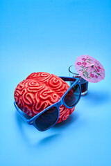 Top view of a red plastic human brain wearing blue sunglasses next to a cocktail party with an umbrella on a blue background.