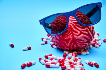 Red rubber brain with sunglasses on surrounded by white and red capsules on a blue surface. Concept of migraines and headache.