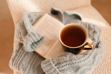 Cup of tea with paper book on knitted sweater in accent chair at home in living room. Good morning.