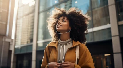 Fototapeten  Horizontal shot of thoughtful curly haired woman uses cellphone for communication over speaker records voice message dressed casually poses against modern city building © Ahtesham