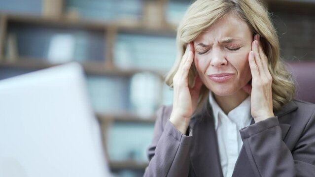 Tired mature businesswoman feeling headache working on computer at workplace in office. Sad overworked woman massages her temples and rubs a sore spot with her hands The female is in pain