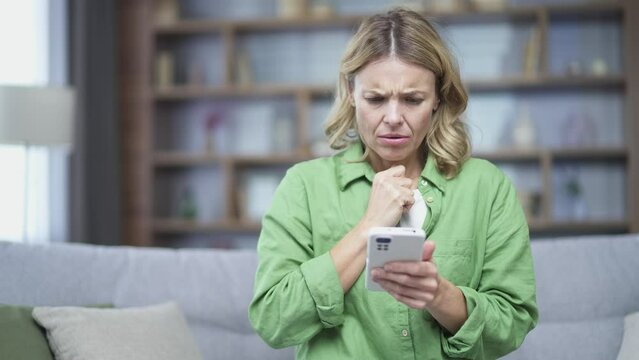 Sad blonde mature woman read bad news on smartphone sitting on sofa at home. Disappointed female received messagen otification with negative response result, worried problem on mobile pone indoor