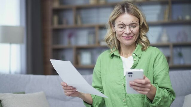 Happy mature woman uses mobile app for spending bills using smartphone phone sitting at home. A female calculates household finances and enters data into a chat bot easy and simple to pay online bank