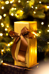 A golden Christmas gift with a golden glittering ribbon in front of a lighted Christmas tree