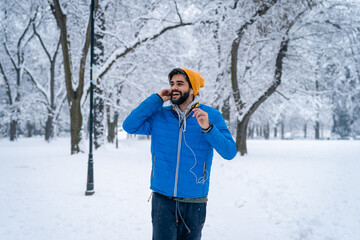 Cheerful young man wearing blue jacket and yellow cap adjusting earphones enjoying music while...